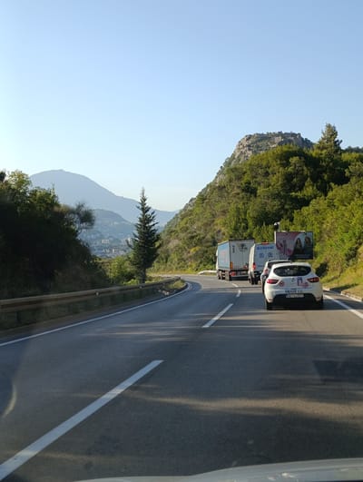 A white car with license plate NK-CU577 is traveling on a road in Zagrađe, Montenegro. The car is following a truck with a white trailer and a white van. There is a billboard on the right side of the road, which advertises "bloomuro". The road is paved and has white lane markings. The area is mountainous with lush green trees. In the background, there is a city with buildings and a large mountain range. The sky is clear and blue.