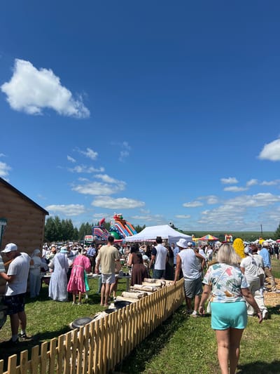 The image depicts an outdoor festival or fair in the city of Kalinino, Russia. A large crowd of people is gathered under a bright blue sky with scattered white clouds. The ground is covered in green grass, and a wooden fence runs across the foreground.
In the background, there are several inflatable attractions, including a large, colorful slide and a bouncy castle. Tents and market stalls are also visible, suggesting a commercial or entertainment event. Some individuals are dressed in traditional or festive attire. The atmosphere appears lively and crowded, indicative of a public gathering. There are trees in the middle ground, and fields extend into the distance under the expansive sky.