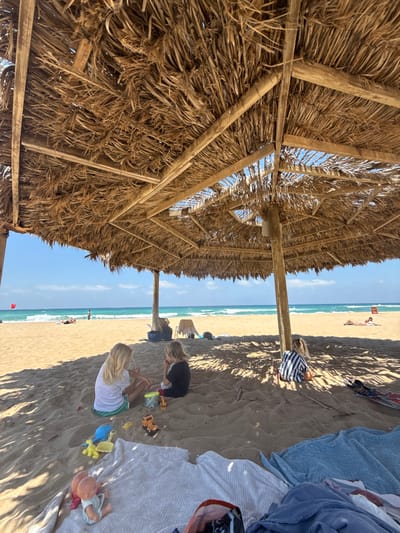 This media file captures a beach scene in Nahariah, Israel, likely during daylight hours under clear skies. The perspective is from beneath a large, thatched umbrella, looking out towards the ocean. The umbrella's natural material and wooden supports are prominently featured in the foreground, casting dappled shadows on the sandy ground.

Several people are present. In the mid-ground, two children are playing in the sand. One child, wearing a black top, is focused on building with toy construction vehicles. Another child, with blonde hair and wearing a white top and green shorts, is also engaged with toys, including a blue shovel and yellow bucket. A doll lies on a blanket nearby. Further out, on the beach, a few other individuals are visible, some lying down and others standing. The turquoise ocean stretches to the horizon, with gentle waves rolling onto the shore.

The overall mood appears relaxed and leisurely, typical of a day at the beach. The presence of the umbrella, towels, and toys suggests a family outing. There is no visible text in the image.