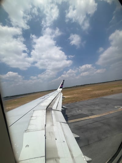 The image, taken from an airplane window, shows an airplane wing extending into the frame. The wing is white with visible panel lines and rivets. The airplane appears to be on a tarmac or runway, with the ground showing patches of dry grass and asphalt. In the distance, there is a clear view of a runway and surrounding landscape. The sky above is blue with scattered white clouds. The location is the city of Apodaca, Mexico, specifically on the grounds of the General Mariano Escobedo International Airport (MTY). The scene depicts a typical moment before takeoff or after landing.