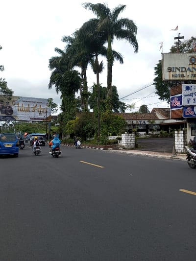 The image displays an outdoor street scene under an overcast sky. A dark asphalt road, marked with a yellow dashed line and a white solid line along a reddish-brown curb, occupies the foreground. Multiple vehicles, primarily motorcycles and scooters, are in motion, traveling away from the viewer. A blue minivan is partially visible on the far left. One motorcyclist in the central foreground is wearing a bright blue and orange poncho.
On the right side of the road, a large, weathered sign for "SINDANGLAYA HOTEL - RESTO" is visible, with smaller signs below, including one partially identifying "S3 Cafe." Two small Indonesian flags (red and white) are affixed to the hotel sign structure. An entrance with white brick pillars leads to the hotel property, which features buildings with tiled roofs largely obscured by dense green foliage.
On the left, a large billboard displays the text "SELAMAT HARI RAYA" (Indonesian/Malay) and features images of individuals. Tall palm trees and various other green trees and bushes are abundant across the midground. Overhead utility wires are visible against the cloudy sky.
Based on the explicit text "SINDANGLAYA HOTEL - RESTO" and "SELAMAT HARI RAYA," along with the presence of Indonesian flags, the scene is located in Indonesia, likely in or near Sindanglaya, West Java, rather than Taohom, Laos.