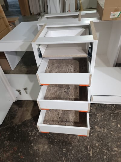 An indoor, eye-level, medium shot shows the assembly of white, laminated particleboard cabinetry with three visible drawers. The cabinetry is partially assembled, with the drawer slides installed on the frame. The drawer fronts are not yet attached. The floor is concrete, with a dark, mottled texture. In the background, there are stacks of what appear to be building materials, including particleboard and cardboard boxes. A box with "FIRMAX" printed on it is visible in the upper right corner. There are no people visible in the image. The overall impression is of a workshop or construction site. The location is not specified in the image.