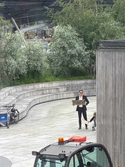 An outdoor, eye-level shot in Kastrup, Denmark, captures a seemingly staged event or protest. The scene is dominated by a paved area with curved, tiered steps and seating, flanked by lush green and silvery-green bushes and trees. In the background, modern architecture with a horizontal wooden slatted facade is visible.
In the mid-ground, a man stands centrally, dressed in a dark suit, white shirt, and sunglasses. He holds up a rectangular cardboard sign that reads, "HANDS OFF MY PRIVATE JET." Behind him, another person is partially visible. Further to the right, a blonde woman is bending over next to a champagne bottle resting on the paved ground, suggesting a celebratory or significant occasion associated with the man's protest.
On the far right foreground, a portion of a grey utility vehicle with an orange flashing light on its roof is visible. To the far left foreground, a bicycle is parked in a rack, featuring a blue basket clearly labeled "SAS," a likely reference to Scandinavian Airlines, aligning with the Kastrup, Denmark location (home to Copenhagen Airport). The time of day appears to be daytime, with diffused light and no strong shadows. The overall impression is one of a public demonstration or performance making a statement about private jet ownership.