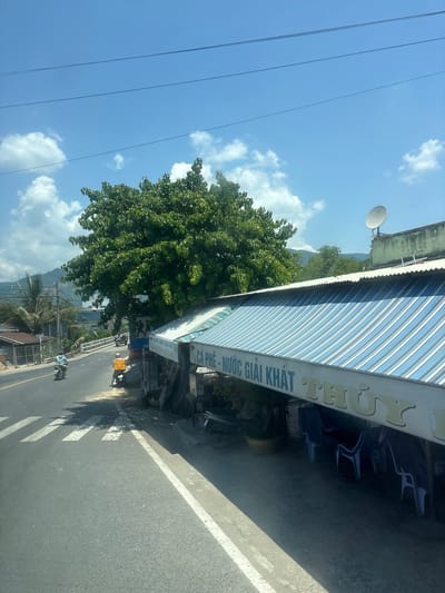 The image depicts a roadside scene in a tropical or subtropical environment. A paved road runs from left to right across the frame, with a pedestrian crossing marked by white stripes in the foreground. Two motorcycles are visible on the road; one in the left lane appears to be moving away from the viewer, while another in the right lane is stationary.
On the right side of the road, a building with a corrugated metal awning extends over a seating area. The awning is striped with shades of blue and white, and a sign in Vietnamese reads "CÀ PHÊ - NƯỚC GIẢI KHÁT THUÝ," indicating it is a coffee and soft drink establishment. Blue plastic chairs are arranged under the awning.
A large, leafy green tree dominates the middle ground, casting shadows on the road and the building. In the background, there are more trees, some structures, a bridge, and a body of water with distant hills. The sky is clear blue with scattered white clouds. A satellite dish is visible on the roof of the building. The overall impression is a casual, open-air establishment along a road in a developing area.