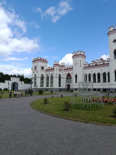 The image depicts the exterior of the Puslowski Palace in Kosava, Belarus. The palace is a white, two-story building with red trim and multiple turrets, designed in a Neo-Gothic architectural style. In front of the palace, a paved pathway leads to a manicured lawn with flower beds and a decorative wire fountain structure.
To the left of the palace, a white wall encloses a courtyard area with a large, open gate. Two elderly individuals are seated on a bench along the pathway in the foreground. Other individuals are visible in the distance, near the palace entrance. The sky is blue with scattered white clouds. The environment is a park or garden setting, characterized by lush green grass, scattered rose bushes, and mature trees in the background.