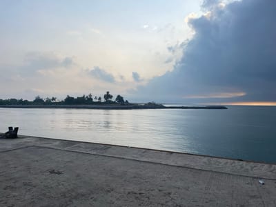 The image depicts a calm ocean scene with a cloudy sky. A long, low island covered in trees and foliage stretches across the center of the frame. A concrete pier occupies the foreground, its surface plain and lightly textured. The water is still, reflecting the sky and island. A breakwater extends into the ocean, protecting the harbor entrance. The sky is dominated by a large, dark cloud on the right, while lighter clouds and a hint of sunlight can be seen on the left.