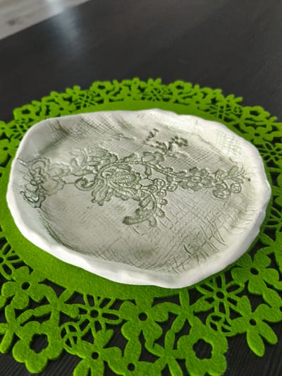 A close-up, high-angle shot shows a small, off-white ceramic dish with a lace pattern pressed into its surface. The dish has an irregular, organic shape with a slightly raised rim. The lace design is in a darker, greenish-grey tone and covers the interior of the dish. The ceramic has a matte finish with visible crackling. The dish rests on a bright green, felt placemat with a cut-out floral pattern. The placemat is on a dark, textured surface, possibly a wooden table. The lighting is soft, creating subtle shadows. There are no people or animals present in the image. The location in the city of Brodi, Latvia, is not discernible from the visual information provided.