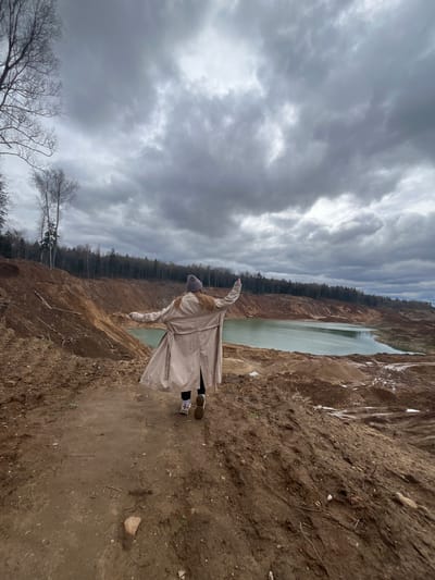 A woman in a light-colored trench coat and beanie stands on the edge of a sandy cliff overlooking a body of water, possibly a quarry. Her arms are outstretched. The sky is overcast with heavy, gray clouds. A line of trees is visible on the opposite side of the water. The ground is dirt and sand. The location is Ivlevo, Russia.
