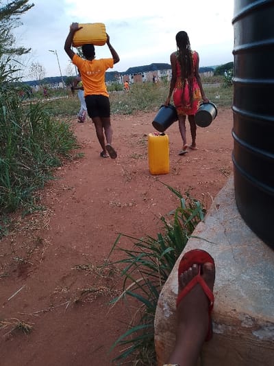 Location: OGBOKO, Nigeria. Two women walk on a dirt path, each carrying water containers. One woman carries a yellow jerrycan on her head, the other carries two black buckets, with another yellow jerrycan placed on the path. The path is bordered by grass and leads towards buildings in the background. A black water tank is visible on the right side of the image. In the foreground, a foot wearing a red sandal rests on a concrete base.