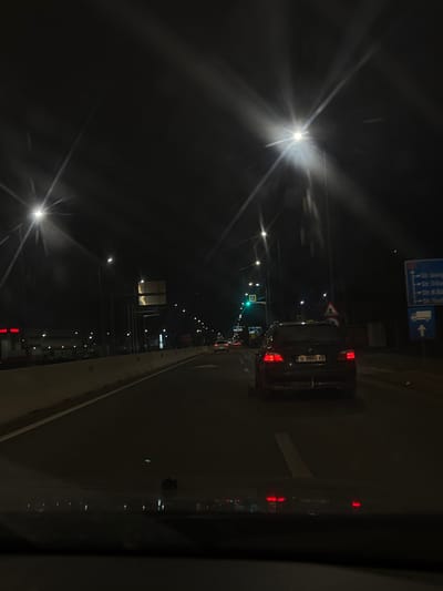 The image displays a nighttime street scene in Jilava, Romania, observed from inside a vehicle. The immediate foreground shows the vehicle's dashboard and a reflection on the windshield. Ahead, a dark-colored BMW sedan, bearing Romanian license plate B 0602 XB, proceeds in the same lane with its red taillights illuminated. The road surface is asphalt, marked by white lane lines. A concrete barrier runs along the left side of the road. Multiple streetlights with distinct radial flares illuminate the environment. Further down the road, a green traffic light is visible, indicating an intersection. On the right, a blue directional sign identifies "Str. Joița," "Str. Toporului," "Str. Teișani," and "Str. Măgurele," with an additional blue sign below indicating a truck symbol with a right-pointing arrow. Distant lights suggest further traffic and structures. No individuals are explicitly visible.