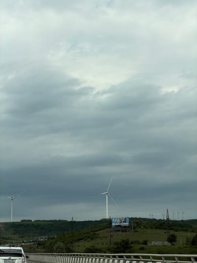 The image shows a landscape in the Tedotsminda area of Georgia, captured from an elevated position, possibly a bridge. In the background, there are two wind turbines alongside a power line. In the middle ground is a green and hilly landscape with a large billboard. In the foreground, part of a vehicle is visible. The sky is overcast.