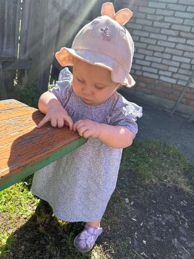 An outdoor, eye-level shot shows a baby girl wearing a light pink sun hat with bunny ears, a grey speckled dress, and purple slip-on shoes. She is leaning over a wooden picnic table, her right hand resting on the table and her left hand near it. The table is weathered with peeling orange paint over green paint. The background includes a grey wooden fence, a brick wall, and sparse grass and dirt. The sun is casting strong shadows across the scene. The location is identified as Novoyeniseyka, Russia.