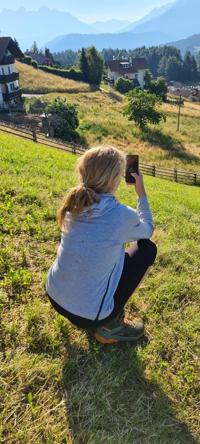 A woman with blonde hair in a ponytail is crouching on a grassy hillside in Deutschnofen - Nova Ponente, Italy, holding a smartphone in her right hand. She is wearing a light blue hooded sweatshirt, black leggings, and green and orange hiking boots. The background features rolling hills with scattered houses and trees, leading to a mountain range in the distance under a clear blue sky. A wooden fence runs along the hillside behind her. The lighting suggests it is late afternoon, with shadows cast on the grass.
