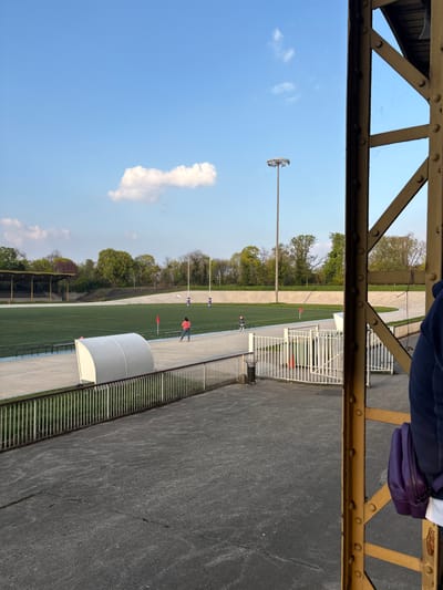 This image depicts an outdoor multi-sport stadium, identified as a facility within Charenton-le-Pont, France. The perspective is from an elevated concrete spectator area, with a yellow metal support beam partially framing the right side of the view. A person's right side, wearing a dark blue jacket and carrying a purple shoulder bag, is visible on the extreme right, adjacent to the support beam.
The foreground consists of a cracked concrete surface. A white metal fence with vertical bars separates this area from a lower concrete perimeter walkway. A black cylindrical trash receptacle is situated near this fence. On the lower walkway, a white, arched team shelter is present. Beyond the walkway is a blue running track, which encircles a central green artificial turf sports field.
Two individuals are on the blue running track: one adult female, dressed in an orange top and blue trousers, stands near the center of the track facing left; and a small child, wearing a light-colored hooded jacket, is positioned further right on the track, facing towards the field. Red flag markers are visible on the perimeter of the green field. Two sets of white rugby goalposts are prominently located on the far side of the field. A white concrete velodrome track surrounds the entire inner sports area, beyond the running track.
In the background, a structure resembling a covered grandstand is partially visible on the far left, positioned behind the velodrome track and partially obscured by trees. A dense line of trees forms the backdrop across the horizon. A tall, slender metal floodlight pole, topped with multiple luminaires, stands on the right side of the field, extending high into the sky. The sky is clear and blue, containing one distinct, elongated white cloud near the center of the frame. The lighting indicates daytime conditions.