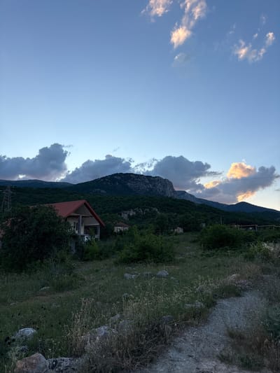 The image depicts a twilight scene in the vicinity of Malyi Mayak, Ukraine. The sky is a gradient of pale blue at the top, transitioning to a darker hue near the horizon. Scattered clouds are present, with some on the right side illuminated by a warm, peachy light, suggesting the sun is setting or has just set.

In the foreground, a rugged, unpaved path runs diagonally from the lower right corner. The ground is covered with dry grass, weeds, and scattered rocks.

Mid-ground features a hillside densely covered with green trees. A house with a prominent red roof and beige walls is visible on the left side, partially obscured by trees. Another smaller building is visible further back amongst the trees. A tall, lattice-like electrical pylon stands to the far left.

The background is dominated by a large, imposing mountain range, its slopes covered in dark green vegetation. The peaks are rugged and rocky. The overall impression is one of a rural or semi-rural mountainous landscape at dusk. No people or animals are visible.