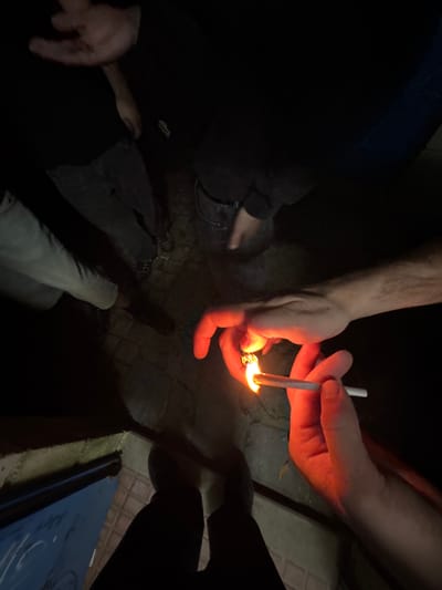 Four people are gathered outdoors at night. One person holds a lit lighter near a cigarette held by another person. The ground is paved. Part of a blue door or panel is visible in the lower right. The location of Amarante, Portugal is not discernible in the image.