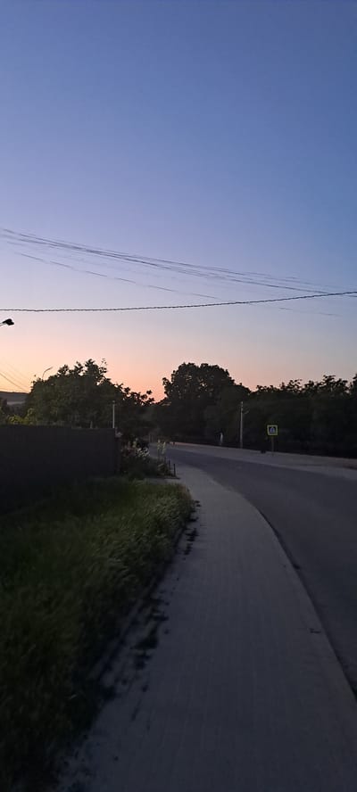 The image depicts a twilight scene on a paved road in Rădoaia, Moldova. The sky transitions from a deep blue at the top to a soft orange and pink hue near the horizon, indicating the sun has recently set or is about to rise. Power lines stretch across the upper portion of the frame. On the left side of the road, there is a dark fence and a strip of grass with tall stalks. The right side features a paved road, with a pedestrian crossing sign visible further down. Trees line the far side of the road. A solitary figure is faintly visible in the distance near a lamppost. The overall atmosphere is calm and quiet, characteristic of the evening in a rural or semi-rural setting.