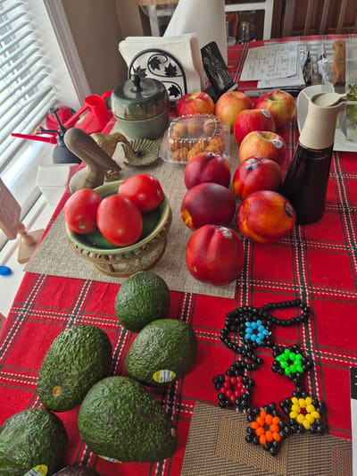 This image shows a still life arrangement of various fruits and decorative objects on a red and green plaid tablecloth. The scene appears to be indoors, possibly on a table near a window with blinds.

Prominently featured are several avocados, located in the lower left and center of the image. To the right of the avocados are a cluster of nectarines and apples. In the upper center, a small green ceramic pot with a lid, resembling an apple, sits near a container of cookies. To the right of the fruit is a dark-colored bottle, likely soy sauce, with a beige ceramic pourer.

Other objects include a small brass pelican figurine, a green decorative dish holding three red tomatoes, and a beaded necklace with flower-like designs in various colors. In the background, a napkin holder with a decorative metal cutout is visible, along with some paperwork and a pen. The overall impression is a collection of items gathered on a table, with a focus on fresh produce and decorative elements. The image does not contain any information about the city of South River, United States of America, or any actions or interactions between people.