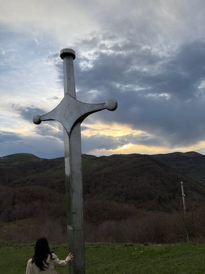 The image shows a large metal sword monument in Zemo Nichbisi, Georgia. The sword is in the foreground, pointing diagonally toward the sky. A mountain range is in the background with sunlight breaking through the clouds. A person with long dark hair and a tan jacket is reaching out toward the base of the sword on the right side of the image. Green grass is visible near the person and the base of the sword. The sky is filled with grey and white clouds, with a section where sunlight is shining through. A wooden cross-like structure is on a hill in the background.