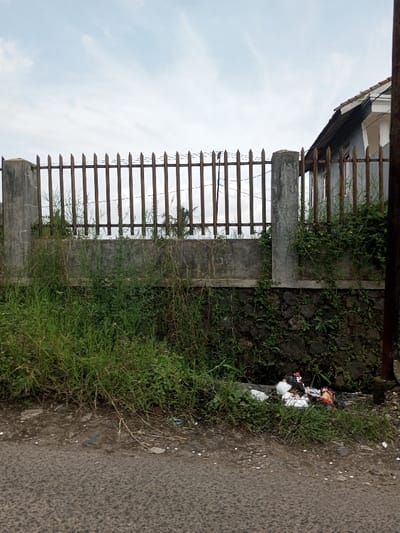 The image displays an outdoor scene in Cibodas, Indonesia, under an overcast sky. In the foreground, a dark asphalt road occupies the bottom portion of the frame. Adjacent to the road, a strip of overgrown green grass and weeds runs along the base of a property boundary. Within this vegetation, on the right side, a pile of assorted refuse, including white plastic and colorful wrappers, is visible.

The boundary itself consists of a multi-tiered structure: a lower stone wall with dark, irregular stones, covered in some green foliage; a concrete wall section above it; and a metal fence with vertical, rust-toned pickets topped with sharpened points. This metal fence is anchored by two concrete posts. Barbed wire is discernible along the upper edge of the metal fence.

Behind the fence, more overgrown vegetation is present, including the fronds of a palm tree. On the far right, a light gray house with a brown tiled roof is partially visible, its side wall aligning with a wooden fence section. A dark vertical pole stands on the extreme right edge of the frame. No individuals or specific actions are depicted within the scene.