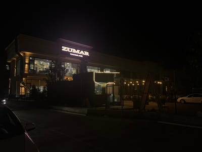 This is a nighttime exterior shot of a restaurant named "ZUMAR TAOMLARI" located in Kyzgaldak, Uzbekistan. The building is lit up, with the restaurant's name prominently displayed in illuminated white lettering above the entrance. The building features large windows that offer glimpses into the restaurant's interior, which is also lit. String lights are draped across an outdoor seating area, casting a warm glow.
The scene is dark, indicating it is nighttime, and the weather appears clear. A white car is parked on the right side of the image, and another car's headlights are visible on the left. A street or road runs in front of the restaurant, and a sidewalk or walkway is visible between the road and the building. The overall mood is calm and inviting, suggesting a pleasant evening dining experience. No people are clearly visible, but the presence of an active restaurant implies patrons and staff are inside or in the outdoor seating area.