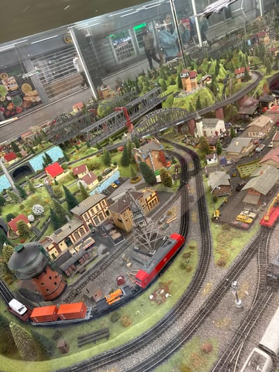 The image shows a detailed miniature diorama of a train layout, likely in Duisburg, Germany, given the branding on the red train engine. The diorama features a complex network of train tracks winding through a landscape populated by small houses, factories, bridges, a water tower, trees, and a river. Several model trains are visible on the tracks, including a red electric locomotive, a train with orange cargo containers, and other freight cars. In the background, through a glass enclosure, a blurred view of what appears to be a shop or market is visible, with some people walking around. The overall scene is meticulously crafted, simulating a realistic miniature environment.