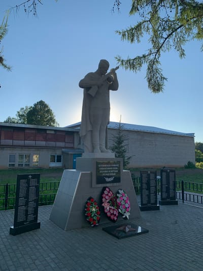 In the city of Svyatitsa, Russia, a monument stands in front of a brick building under a clear blue sky. The monument features a statue of a soldier holding a bugle. Below the statue, a central plaque reads, "LET'S REMEMBER ALL OUR FALLEN. IT IS NOT NECESSARY FOR THE DEAD - IT IS NECESSARY FOR THE LIVING."
To the left of the central plaque, there are two black granite slabs with lists of names. To the right, two more similar slabs are visible. In front of the monument, two floral wreaths are placed on the ground, one with pink and white flowers, and the other with red and pink flowers. A red star is also visible on the ground in front of the wreaths. The area around the monument is paved with light-colored square bricks. A dark metal fence encloses the monument area. The background shows a long, low-rise building with a light-colored roof. Trees are visible behind the building and to the right. A branch of a coniferous tree hangs over the top left of the image.
