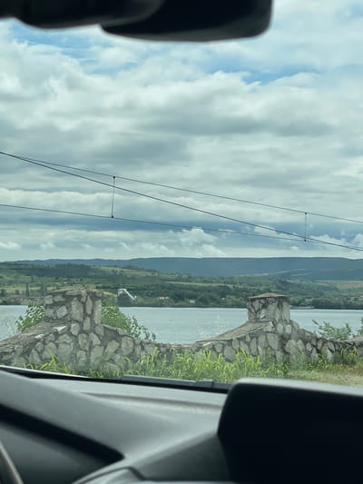 This image captures a wide landscape view, seemingly taken from inside a vehicle looking out through a window. The scene is dominated by a large body of water, distant hills, and a cloudy sky.
In the foreground, elements of a car's interior are visible on both the left and right edges, in dark grey and black tones, framing the picturesque outdoor scene. These include parts of the door frame and window trim, suggesting the photograph was taken from a passenger's perspective.
Immediately beyond the car's interior, a rugged stone wall or embankment runs along the edge of a wide expanse of water, which is light blue-grey and reflects the cloudy sky. Green foliage, including grass and small bushes, grows on and behind this stone structure. A small, light-colored, angular structure, possibly a pier or a small utility building, is visible at the water's edge in the midground.
Stretching across the horizon in the background is a long chain of dark green hills or mountains, appearing to be covered in trees. Above this landscape, the sky is largely filled with white and grey cumulus clouds, with some lighter blue areas visible. Several thin, black utility lines (likely power lines) are strung horizontally across the upper portion of the sky.
The image depicts a serene daytime setting under a cloudy sky, suggesting moderate weather conditions. The presence of a wide body of water and distant hills is consistent with the provided location context of Drobeta-Turnu Severin, Romania, suggesting the water is likely the Danube River. There is no visible text in the image.
