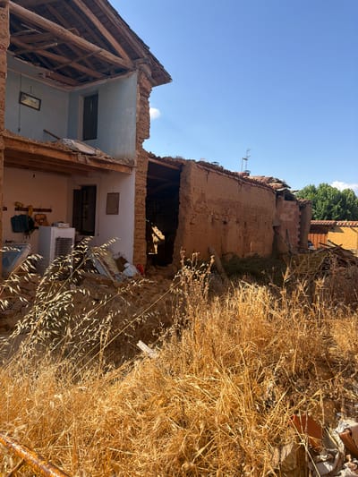 A side view of a damaged, traditional Spanish house in Arcahueja, Spain, under a clear blue sky. The building's adobe walls are crumbling, and the roof has partially collapsed, exposing wooden beams. Debris, including what appears to be a refrigerator and various household items, is scattered around the structure, partially obscured by dry, golden grass in the foreground. The scene depicts a state of disrepair, suggesting it may have been abandoned or damaged by an event. The surrounding environment is rural, with greenery visible in the background.