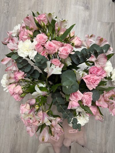 An overhead, close-up shot captures a vibrant floral bouquet resting on a light gray wooden floor. The bouquet is primarily composed of various shades of pink, white, and green.
The main flowers visible are numerous pink roses, ranging from tight buds to partially open blooms, providing a lush, romantic feel. Interspersed among the roses are delicate pink and white alstroemeria (Peruvian lilies), characterized by their striped petals and often speckled centers. Fluffy, ruffled white flowers, resembling carnations or similar blossoms, add volume and brightness to the arrangement. Dark green, rounded eucalyptus leaves are strategically placed throughout the bouquet, offering a contrasting texture and a touch of deep green amidst the softer colors.
The background is a light gray wooden floor with distinct plank lines and a visible wood grain pattern. To the right side of the image, peeking out from behind the flowers, is a soft, light pink, furry or plush object, possibly a slipper or part of a cozy garment. A small tag on this pink object is visible, displaying some dark text that appears to read "R. MATTHEW".
The lighting is soft and even, highlighting the colors and textures of the flowers. The bouquet appears fresh and full, though some of the alstroemeria and lower rose petals in the bottom right section show slight signs of wilting. The overall impression is one of beauty and tranquility.
