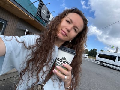 A woman with long, curly brown hair takes a selfie outdoors. She is wearing a light blue t-shirt and holding a coffee cup with Russian text that reads "КОФЕ на высоте" which translates to "Coffee at altitude." The background features a building with a green roof and a sign with the text "ДАРЮ АЛТАЙ" which translates to "Gift of Altai." In the distance, a white van is visible on a road, and the sky is cloudy with patches of blue. The location is identified as T/B Seminskii Pereval, Russia.
