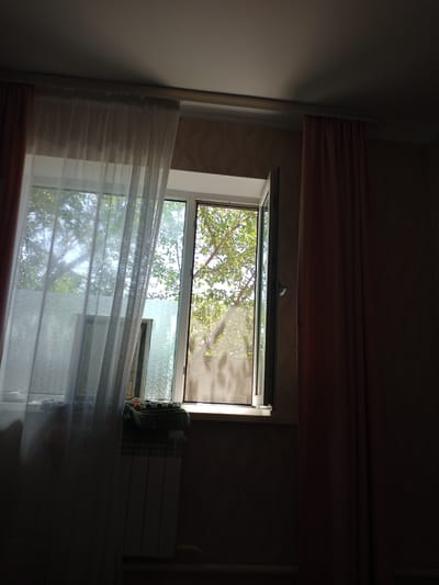 The image depicts an interior view of a room with a large window that is partially open. Sunlight streams through the window, illuminating the room and casting shadows. Lush green trees and foliage are visible outside the window. The window is adorned with sheer white curtains on the left and heavier pinkish-brown curtains on the right. On the windowsill, there are decorative items and a radiator is positioned below the window. The overall scene suggests a quiet, possibly residential, setting in Kuntuu, Kyrgyzstan, where the natural light plays a significant role in the ambiance of the space.