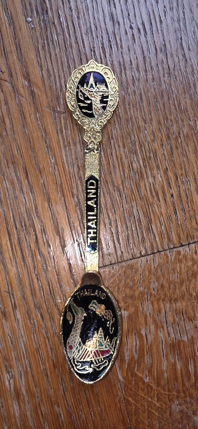 A gold-toned souvenir spoon with black enamel detailing rests on a wooden surface. The spoon's handle is inscribed with the word "THAILAND" in black, capitalized lettering. The top of the spoon features a circular emblem with a gold-toned ornate border, enclosing a black background with an image of a Thai temple or palace rendered in gold and purple. The bowl of the spoon is oval and also features a black background. On the bowl, there is an image of a mythical creature, possibly a Naga or dragon, depicted in gold with red accents, alongside a miniature depiction of Thai architecture, complete with colorful spires and flags. The wooden surface appears to be made of planks with a prominent grain pattern. The image is taken from directly above, with the spoon lying flat.