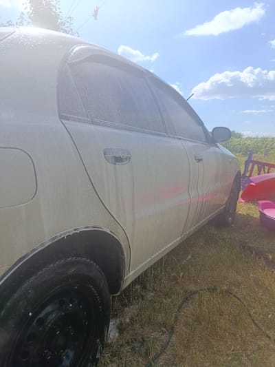A light-colored sedan is being washed outdoors on a sunny day. The car's exterior is covered in white foam and water droplets, indicating it is being sprayed with water and soap. The vehicle is parked on a patch of dry grass, with a black hose visible on the ground. In the background, a fence line and some greenery suggest a rural or suburban setting, potentially in the village of Pishchane, Ukraine. Partially visible to the right are children's toys, including a purple and pink tub and a brightly colored chair, suggesting a family environment. The sky above is blue with scattered white clouds, and sunlight is reflecting off the car's wet surface.
