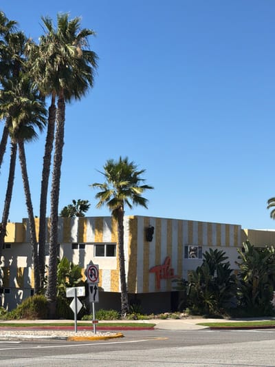 The image depicts the exterior of a two-story commercial building located at 20040 S Western Ave in Torrance, California, United States. The building's facade is distinctive, featuring vertical stripes of yellow and off-white mosaic tiles. A prominent, stylized red sign spelling "Tile" is affixed to the mosaic facade on the right side of the main section. Above this sign, a dark, vertical rectangular element is integrated into the tiled pattern. Several rectangular windows with white frames are visible on both the lower and upper levels of the building.
In the foreground, a multi-lane asphalt road is present, separated from the viewer's perspective by a raised concrete median with yellow curb paint. A cluster of traffic signs on a single pole is positioned on this median. From top to bottom, the signs include a red circular "No U-turn" symbol, a white rectangular sign with black arrows indicating "straight or right turn only," and a smaller, less distinct white rectangular sign below.
Numerous palm trees, varying in height and species, dominate the left side of the frame and are scattered across the scene. A group of tall, slender palm trees stands in front of the building on the left, casting shadows onto the facade. A shorter, fuller palm tree stands directly in front of the mosaic-tiled section of the building. Lush green landscaping, including broad-leafed plants, partially obscures the lower portion of the building and lines the perimeter of the property and the median. The sky is clear and brilliant blue, occupying the upper half of the image, indicating a sunny day. No individuals or vehicles are visible in the scene.