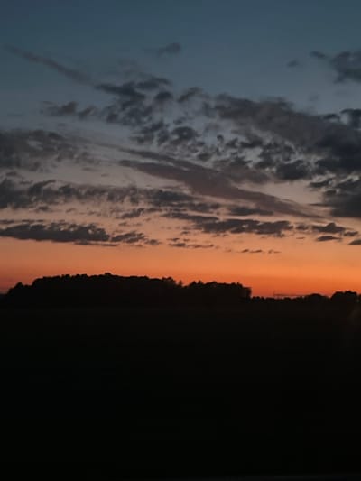 The image depicts a silhouetted landscape at dusk. A dark, treed horizon line cuts across the lower third of the frame. Above this, a vibrant band of orange transitions into a deep blue sky filled with scattered, wispy clouds. The scene is devoid of any human presence or identifiable structures, suggesting a rural or natural setting. The location is specified as Поварёнки, Russia.