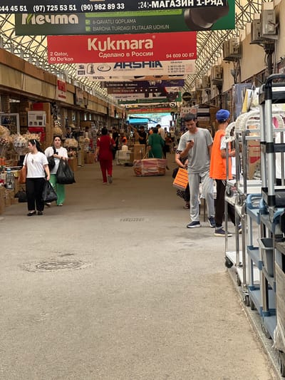 The image shows a bustling marketplace scene in Eshanguzar, Uzbekistan. In the foreground, a man in a grey t-shirt and pants is looking at his phone, while a person in an orange vest stands next to him. To the left, two women are walking away from the camera, each carrying shopping bags. One woman is wearing a white t-shirt and black pants, and the other is wearing a white t-shirt and green pants. In the background, various stalls and shops are visible, with goods displayed and people shopping. Overhead banners with text in Uzbek and Russian are prominent. The ground is paved, and a manhole cover is visible in the lower left quadrant of the image. Several shopping carts are lined up along the right side of the frame. The overall impression is one of a busy commercial area.
