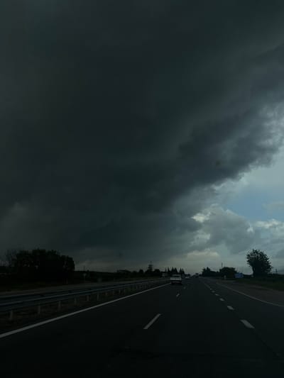 The image depicts a highway adjacent to what appears to be a rural area in Zlatina, Bulgaria. The highway stretches into the distance, with a metal guardrail visible on the side. Several cars are visible on the road. The sky is dominated by dark, ominous storm clouds, with a small patch of lighter clouds visible.