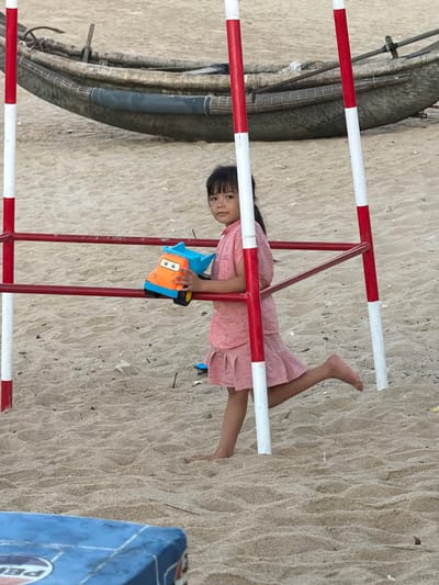 A young Asian girl wearing a pink dress is hanging upside down from a red and white striped metal structure on a sandy surface. She is holding a blue and orange toy car. The location appears to be a beach or sandy area in Thuận An Ward, Vietnam, given the presence of sand and a glimpse of a blue object possibly related to the beach environment. There are no other people visible in the frame.