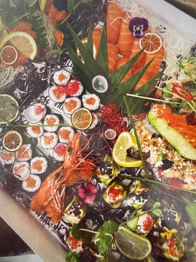 A close-up overhead shot shows a sushi platter. The platter features an assortment of sushi rolls with salmon and red roe, as well as slices of salmon sashimi. Garnishes include slices of lemon, lime, and dried orange, along with a large cooked prawn, red coral-like pieces, and edible flowers. The platter is laid out on a surface with a black and white patterned paper liner. The overall presentation is elaborate and decorative.