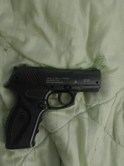 A black CO2-powered air pistol, identified by the markings "C11" and "CBC inc. Fl, USA, cal. .177 (4.5mm) Warning! Not a Toy. Read manual before use.", is resting on a light green quilted fabric. The pistol is positioned horizontally, with the barrel pointing towards the right side of the frame. The fabric has a subtle pattern of bamboo leaves printed on it. The scene is indoors, likely in a residential setting in Sheremet'yevka, Russia. There are no individuals present in the image. The lighting appears to be ambient, with no specific light sources clearly visible.