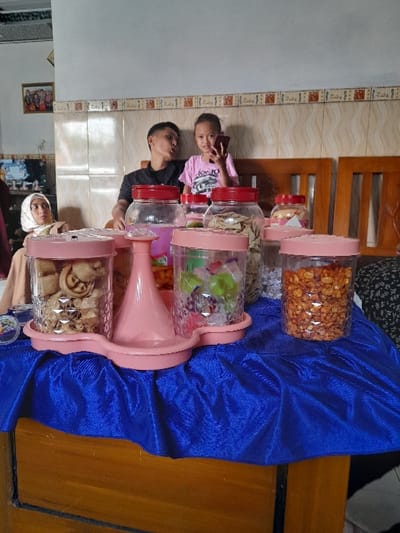 The scene is a living room in a home in Kecamatan Purwoasri, Indonesia, located at coordinates -7.637 latitude and 112.141 longitude. It is a brightly lit room with white walls and a patterned tile floor. There are three people present: a man in a blue shirt, a young girl in a pink shirt and a woman in a white hijab. The man is sitting on the couch with his back to the camera, the girl is seated next to him and the woman is standing behind the man. The woman is looking at the camera.  The room is decorated with a blue tablecloth, a wooden table, and numerous glass jars of snacks. The jars are arranged on the tablecloth. The scene appears to be a family gathering. The woman is likely taking a picture of the man and the girl.  There are a few pictures on the wall behind the couch.  The light in the room is bright and warm. The air is still, and the room is clean and tidy.  There is no furniture other than a couch, a wooden table, and a blue tablecloth. The jars contain snacks, which are likely for a gathering. The people are dressed casually.  The scene is a warm and inviting home environment.