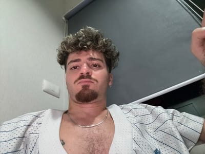 This is a first-person perspective, upside-down image of a person. The subject has curly, light brown hair and a goatee. They are wearing a white jersey with thin blue pinstripes and a perforated texture. A silver chain is visible around their neck, and what appears to be a tattoo is on their chest. The background is predominantly a flat gray surface, likely a desk or table, with a white wall and a light switch visible on the right. The lighting suggests an indoor setting, possibly during the day. There are no clear indicators of weather or a specific event. The expression on the subject's face is neutral.