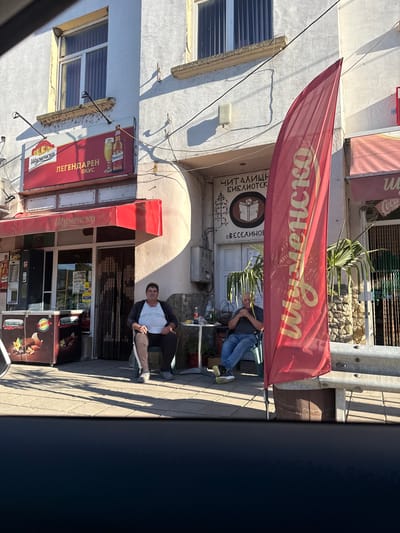 This image depicts the exterior of a commercial establishment in the city of Veselinovo, Bulgaria. In the foreground, a large red flag with yellow lettering, likely advertising a brand, stands prominently. Two men are seated on chairs outside the building. The man on the left is wearing a light-colored t-shirt, dark pants with a white stripe, and a dark jacket. He is looking towards the camera. The man on the right is wearing a dark t-shirt and blue jeans, and is also seated. In front of the building, there is an ice cream freezer. Above the entrance, a sign displays text in Cyrillic, and to the left of that, another sign features a beer bottle and glass. The building itself appears to be painted white and has multiple windows. The ground is paved with interlocking bricks. The overall scene suggests a casual daytime setting outside a shop or cafe.