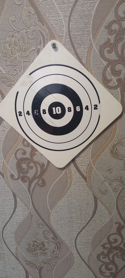 The image shows a dartboard hanging on a wall with patterned wallpaper. The dartboard is diamond-shaped and white, with concentric black circles and numerical scoring. The numbers visible on the dartboard are 2, 4, 8, 10, 8, 6, 4, and 2. The wallpaper has a textured pattern with floral motifs and wavy lines in shades of beige and brown. A single screw is visible securing the dartboard to the wall. The scene appears to be indoors. The location is identified as Идрисово, Russia.