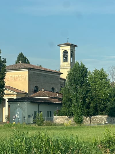 The image captures a view of a church in Pumenengo, Italy, on a clear day. The church features a prominent bell tower with a cross at its apex, several bells visible within its arches, and a tiled roof. The main church building is constructed with stone and yellow stucco, adorned with a fresco depicting figures in a frieze along the upper facade. Adjacent to the main structure is a smaller, lighter-colored building with arched doorways and windows. The church is partially obscured by lush green trees and foliage. In the foreground, tall green grass and reeds are visible, suggesting a rural or park-like setting. A stone wall runs along the property line, and a road is visible behind it. Various signs are present, including a speed limit sign indicating "30." The sky is a clear, pale blue.