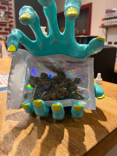 An indoor scene in Clermont-Ferrand, France, displays a light blue, stylized five-fingered object with yellow fingertips holding a clear, iridescent, resealable plastic bag. The bag contains an irregular mass of dark green, dried plant matter. The entire arrangement rests on a light brown wooden surface. The blurred background reveals a light-colored brick wall section to the left, and shelves stocked with numerous small bottles, likely e-liquid or similar products, to the right, suggesting a commercial or retail setting. A white plastic cap is visible on the wooden surface to the right of the primary subject. No individuals are present.