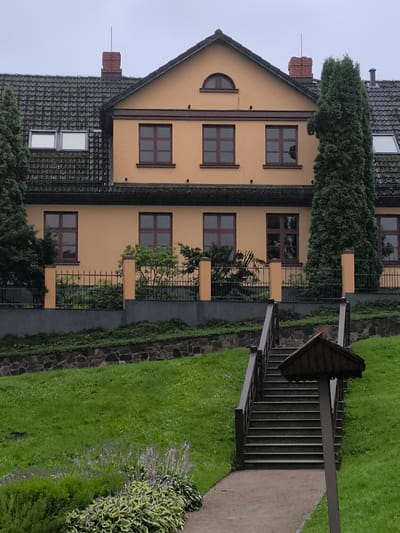 This is a vertical, eye-level, outdoor, full shot of the facade of a large, multi-story, light yellow house with dark brown window frames and a dark gray tiled roof. The house is set back from the viewer, with a steep grassy slope in the foreground leading up to a set of dark brown wooden stairs that ascend to the house. The stairs are flanked by green grass, and at the top of the stairs is a small, dark brown, roofed structure on a pole. A dark metal fence runs along the front of the house, behind which are various bushes and trees, including a tall, dark green evergreen tree to the right of the house and another to the left. Two skylights are visible on the left side of the roof, and a brick chimney with a lightning rod is on the peak of the roof between the two sides. The sky is overcast and a dull gray. This is likely a property located in the city of Kuldīga, Latvia, possibly in a residential area.