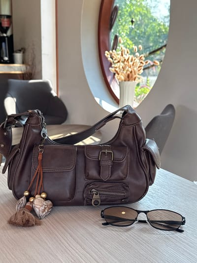 The image depicts an interior scene, likely a cafe, located in San Phra Net, Thailand. On a light-colored wooden table, a dark brown, multi-pocketed shoulder bag is centrally positioned. The bag features two flap pockets with buckle closures on the front, a smaller zippered pocket below, and an additional side pocket. A decorative charm, consisting of brown tassels, gold beads, and two heart-shaped, marbled ornaments, hangs from the bag's strap attachment. To the right of the bag, a pair of black-framed sunglasses with amber-tinted lenses lies flat on the table.
In the background, a distinctive oval-shaped window with a dark brown frame provides natural illumination, revealing green foliage outdoors under bright daylight. A white vase containing dried ornamental grasses is placed on the window ledge. To the left of the window, a dark gray upholstered chair is visible, with a dark object (possibly a coffee machine) and a white container on a shelf further in the background. A second dark gray chair is partially visible on the right side of the frame, behind the table. No individuals are present in the scene.