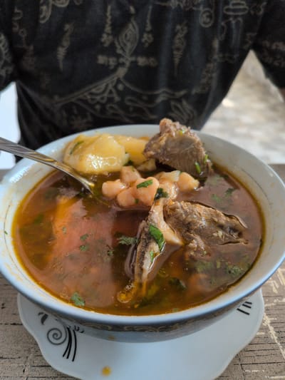 A close-up shot shows a white bowl filled with a hearty meat and chickpea stew. The stew is a rich, reddish-brown color, with chunks of tender meat, visible bone fragments, and a generous portion of chickpeas. Chopped green herbs, likely parsley or cilantro, are sprinkled throughout the dish, adding a vibrant contrast to the darker ingredients. Pieces of yellow potato are also visible beneath the surface of the broth. A spoon, partially submerged in the stew, rests within the bowl. The bowl itself has a white interior and a rim with a subtle dark pattern. It sits on a decorative white saucer with a spiral design.
The background on the left is a dark, textured fabric with a faint, intricate pattern, possibly clothing. The background on the right is a rough, textured surface, likely a wooden table or countertop, with a light brown hue. The lighting suggests it's daytime, and the overall setting appears casual, possibly a meal being enjoyed at home or in a local eatery. There is no visible text in the image. The food appears to be a traditional Azerbaijani dish, fitting for a meal in Shaki, Azerbaijan, given the context. The image focuses on the appetizing presentation of the stew, highlighting its rich ingredients and appetizing appearance.